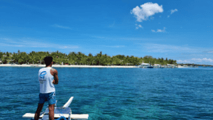 man in white vest at front of boat approaching a tropical island with white sandy beach and palm trees